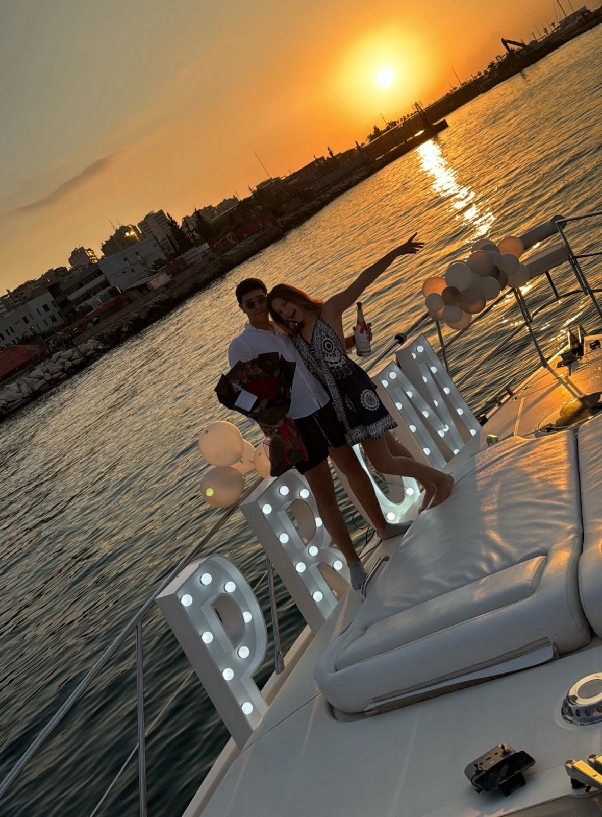 Couple celebrating prom on a luxury yacht at sunset, holding flowers and champagne with illuminated letters and a glowing sea backdrop.